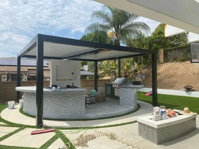 Freestanding Four Seasons louvered roof with black frame over a curved outdoor kitchen and built-in grill